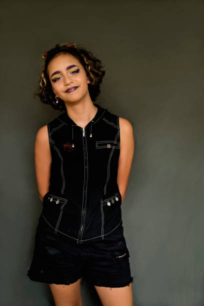 Portrait of a mixed-race non-binary teenager in studio. They have curly brown hair with blonde streak and are wearing a sleeveless black top with DIY embroidery on it. Make-up is in the coolers of the Non-Binary LGBTQ+ flag. Vertical waist up studio shot on khaki background.