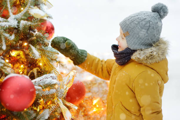 little boy admiring artificial christmas tree decorated with gold and red balls and flowers. traditional city outdoors christmas market. urban celebrate decoration - balloon child winter snow imagens e fotografias de stock