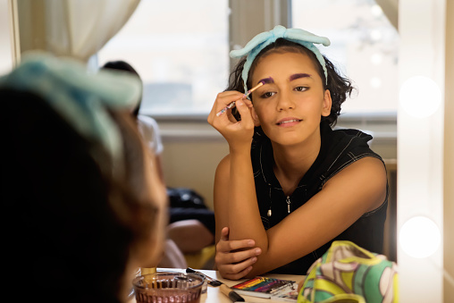 Portrait of a mixed-race non-binary teenager in make-up session in bedroom. They have curly brown hair with blonde streak and are wearing a sleeveless black top with DIY embroidery on it. Supportive mother in the background is wearing a Straight Ally LGBTQ+ t-shirt. Horizontal waist up indoors shot with copy space.
