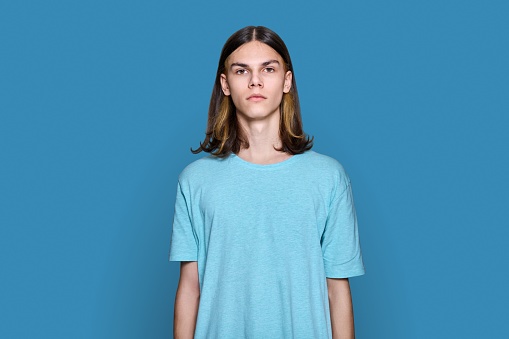 Portrait of serious teenage guy looking at camera on blue color background. Handsome young male with long hair hairstyle posing in studio. Youth, students, people concept