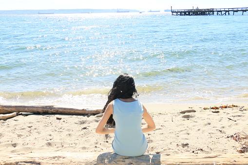 An Asian, Indian woman sitting on a beach looking out at the Pacific Ocean and a pier. She is wearing a blue sleeveless dress.