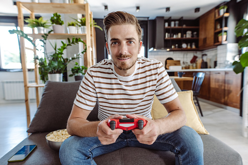 Happy young gamer holding a joystick and enjoying playing video games at home. He is sitting on the sofa and relaxing