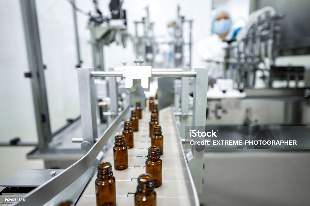 Two female employees in a pharmaceutical laboratory seen while controlling the process of drugs manufacturing Small brown bottles seen perfectly arranged in a laboratory machine during manufacturing in a pharmaceutical factory while two female employees in a protective clothing are controlling the process. Probiotic Stock Photo