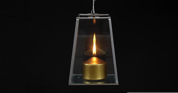 In the dark, a golden candle burns in a lamp. The flame of a beautiful candle is reflected in the glass walls of the lantern. Close-up shot of a lantern with a candle on a black background.