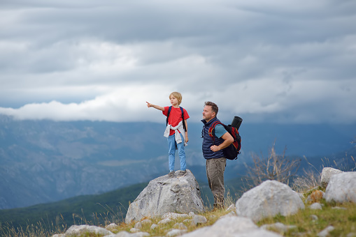 Cute schoolchild and his mature father hiking together on mountain and exploring nature. Child watching a clouds. Concepts of adventure, scouting and hiking tourism for family with kids.