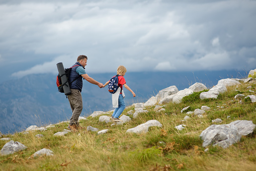 Cute schoolchild and his mature father hiking together on mountain and exploring nature. Concepts of adventure, scouting and hiking tourism for family with kids.