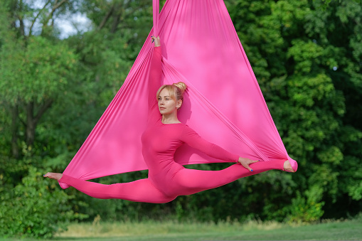 Beautiful young woman practicing aerial yoga and sitting on a split at the tree hang in park. Woman doing antigravity yoga with the use of a hammock.