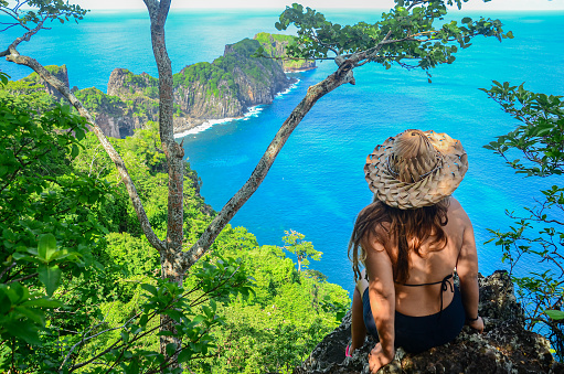 A young woman sitting on Ponta da Sapata Viewpoint and wearing straw hat. Capim açu trail, Fernando de Noronha archipelago, Brazil