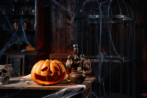 Halloween. Scary Halloween pumpkin with carved face on table in dark room with candles, spider web, and cage on background. Copy space for text. Spooky atmosphere. Mysterious room in dark dungeon