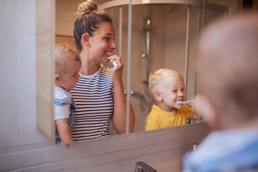 Cheerful woman holds one son in her arms, while she teaches the other to brush his teeth by himself