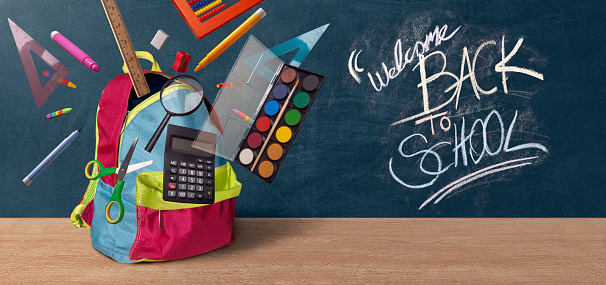 School stationery set floats around a colorful school backpack placed on wooden desk. Welcome back to school text written with colorful chalks visible on the chalkboard
