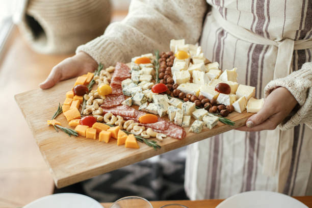 Woman holding cheese board on background of stylish christmas table with fir branches and candles. Cheese appetizers and salami in shape of christmas tree, creative food arrangement for holidays stock photo