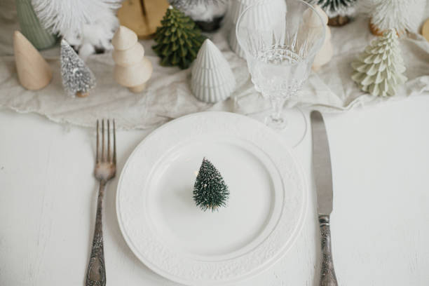 istock Stylish Christmas table setting flat lay. Modern little christmas tree on plate, vintage cutlery, wineglass, modern christmas trees and houses on white rustic table. Holiday brunch 1424143330