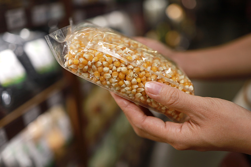 Young Asian woman doing grocery shopping in a supermarket, close up of her hand choosing a pack of organic whole corn. Healthy eating lifestyle.