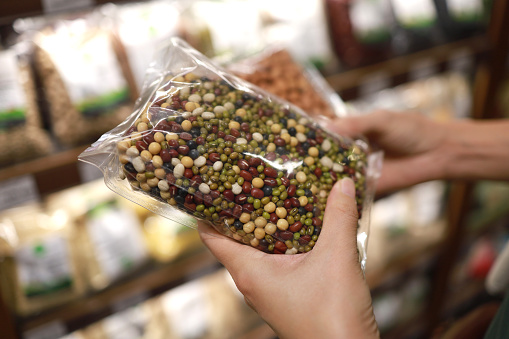 Young Asian woman doing grocery shopping in a supermarket, close up of her hand choosing a pack of organic mixed beans. Healthy eating lifestyle.