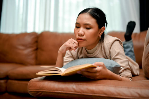 istock Relaxed and chill Asian female reading a book or novel while laying on her comfy leather sofa 1424152141