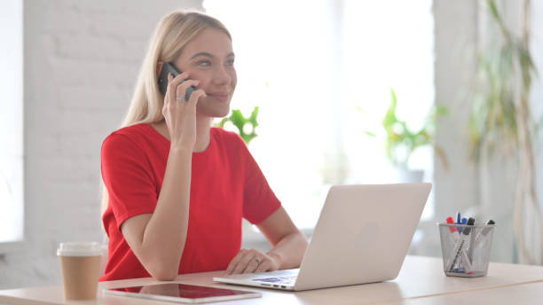 istock Young Blonde Woman Talking on Phone while using Laptop in Office 1424162685