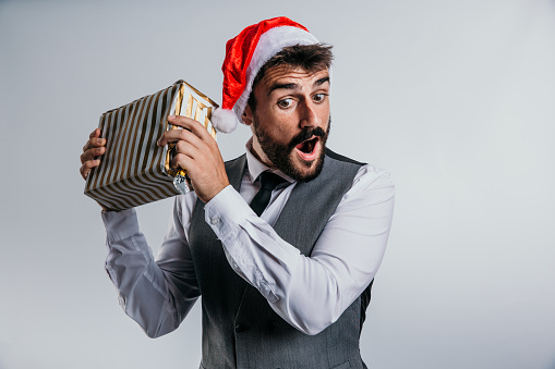 Elegantly suited, bearded, young adult male, with a Santa hat making fun faces while being playful with a wrapped gift, guessing what's in. New Years concept. Studio white background