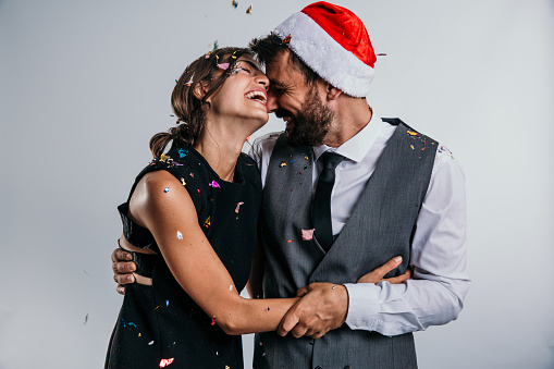 Beautiful brunet wearing elegant dress hugging her handsome bearded partner in a gray suit, for a New Years party with a confetti flying above them. Standing isolated in a white background studio