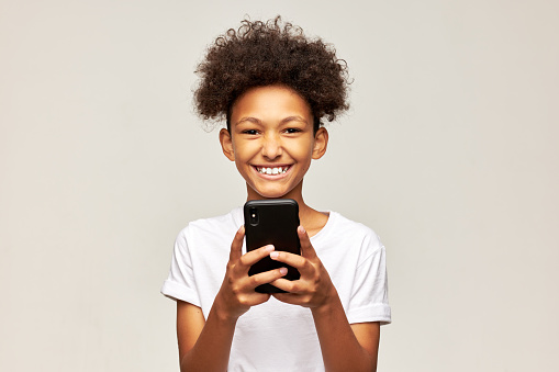 Handsome african american kid with afro hairstyle using mobile app on his smartphone to learn foreign language isolated on gray studio background with happy cheerful facial expression