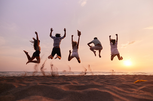 Rear view of playful friends having fun while jumping with raised arms on the beach at sunset. Copy space.