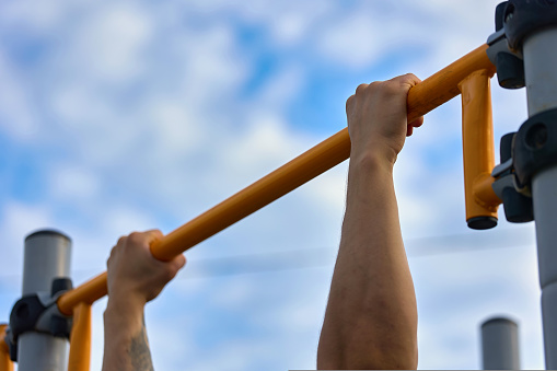 Close-up of the athlete's hand hanging on the horizontal bar against the blue sky. A strong athlete performs an exercise at the turnstile for pull-ups. Sports on the crossbar