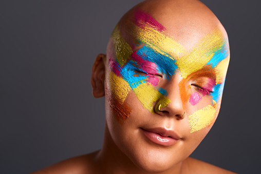 Face, paint and art with a bald woman in studio against a gray background with color on her skin. Creative, cosmetic and artistic with a young female posing eyes closed for freedom and expression