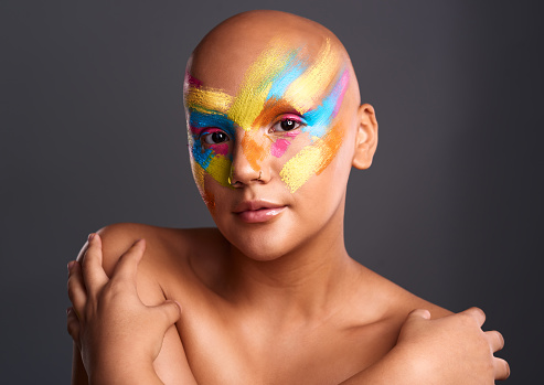 Beauty, face paint and portrait of a beautiful woman with colorful bodypaint and artistic makeup against a grey studio background. Fine art, creative look and skin of confident and bald female model