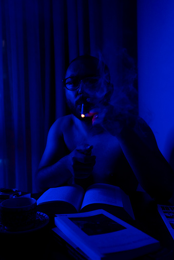A man smokes while working at a desk in a dim blue light environment