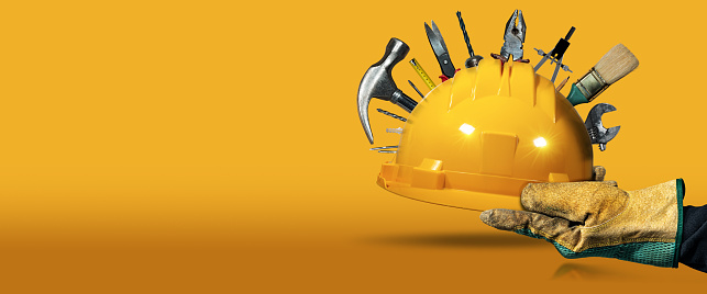 Manual worker with protective work gloves holding a yellow safety helmet with many work tools, on a yellow and orange background with copy space and reflections.
