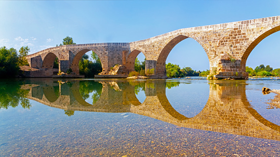 Seljuk Bridge in Aspendos. The Eurymedon Bridge. Crooked bridge, reflected in calm water. An ancient building across the Kopruchay river. Antalya region, Turkey.