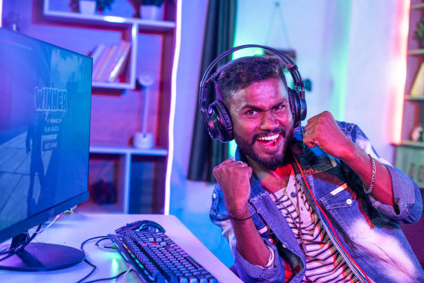 istock Cheerful gamer with headphones celebrating game by shouting raising hands while playing online video game on computer by looking at camera at home - concept of achievement, tournament and happiness 1424185432