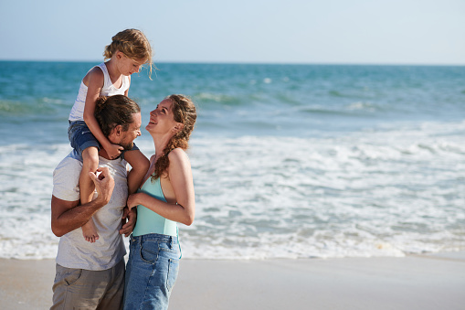 Happy family of three standing on beach, father carrying daughter on shoulder and looking at wife