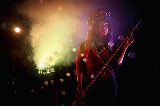 Woman magician illusionist of circus with cane showing soap bubbles show at black background. Female actress in theatrical clothes with stylish hat in stage costume. Concept of theatre performance