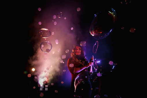 Woman magician illusionist of circus showing soap bubbles show at black background. Female actress in theatrical clothes with stylish hat in stage costume. Concept of theatre performance. Copy space