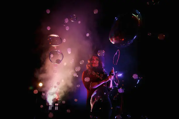 Photo of Woman magician illusionist of circus showing soap bubbles show at black background. Female actress in theatrical clothes with stylish hat in stage costume. Concept of theatre performance. Copy space