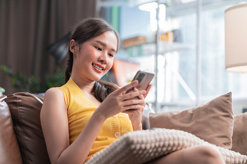 attractive young asia adult woman using a mobile phone while relaxing on the sofa at home,asian female using smartphone surfing the internet social media or chating with her friend smiling cheerful moment at home