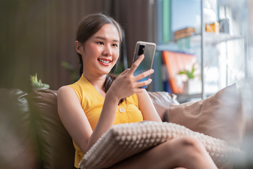 attractive young asia adult woman using a mobile phone while relaxing on the sofa at home,asian female using smartphone surfing the internet social media or chating with her friend smiling cheerful moment at home