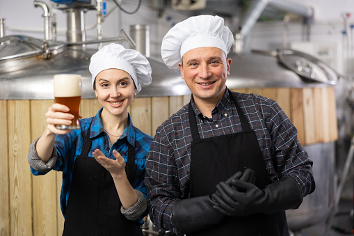Smiling man and woman brewers presenting new beer in beer factory. Woman making thumb up gesture.