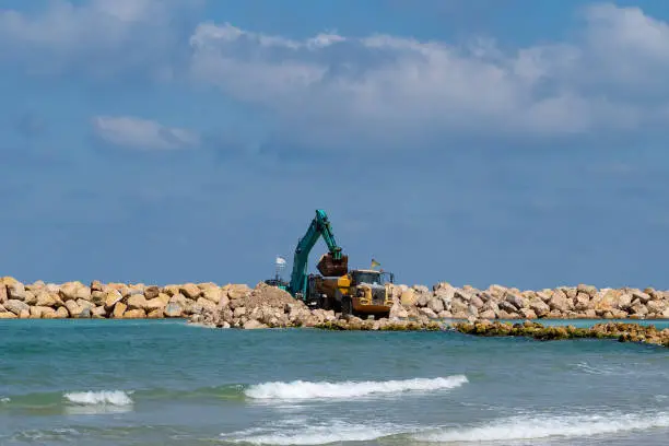 An excavator and a mining truck move stones into the sea. Construction of breakwaters to protect the beach in Netanya in Israel. Industrial building equipment and machines.