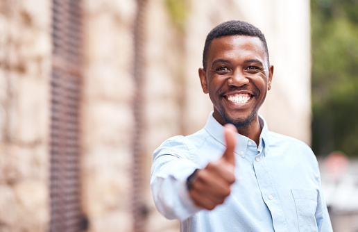 Portrait of an African man with a smile and thumbs up standing in the outdoor urban city street. Happy, excited and positive black guy with a like or agreement gesture outside a building in the road.