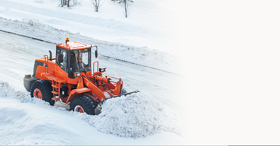 A large orange tractor removes snow from the road and clears the sidewalk. Cleaning and clearing roads in the city from snow in winter. Snow removal after snowfalls and blizzards. banner