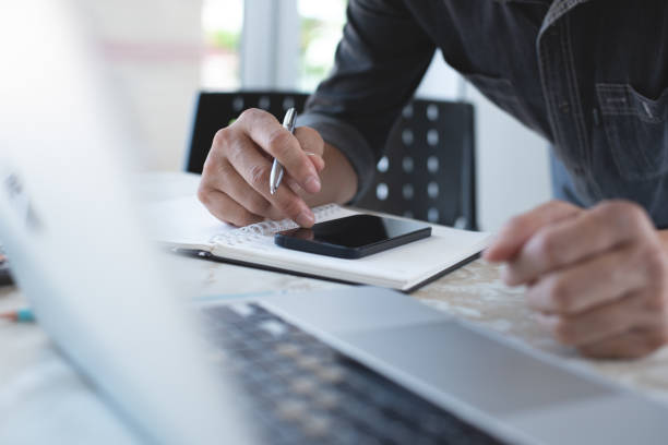 istock Close up of casual business man using mobile smart phone, surfing the internet, searching information and working on laptop computer with notepad on office table 1424205708