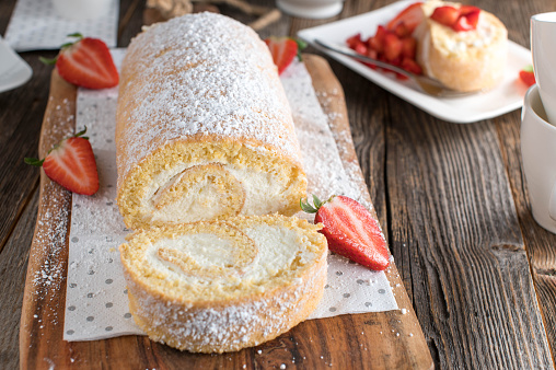 Delicious cake dessert with a homemade fresh baked swiss roll or biscuit roll. Filled with whipped cream and served with marinated strawberries on wooden table