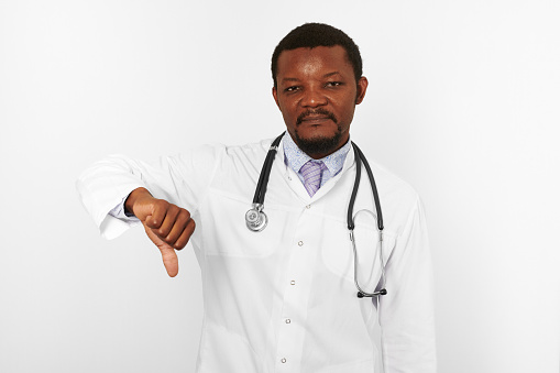 Unhappy black bearded doctor man in white robe with stethoscope shows dislike gesture, isolated on white background. Sad adult black african american physician therapist portrait diagnosed disease