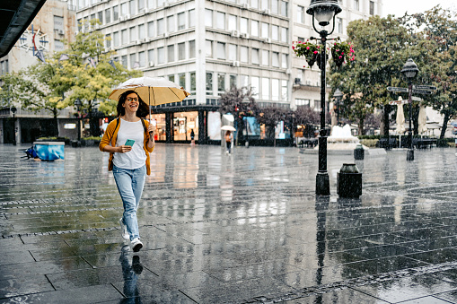 Beautiful young woman running down the street on a rainy day, holding an umbrella and a smart phone.