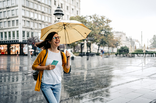Beautiful young woman running down the street on a rainy day, holding an umbrella and a smart phone.