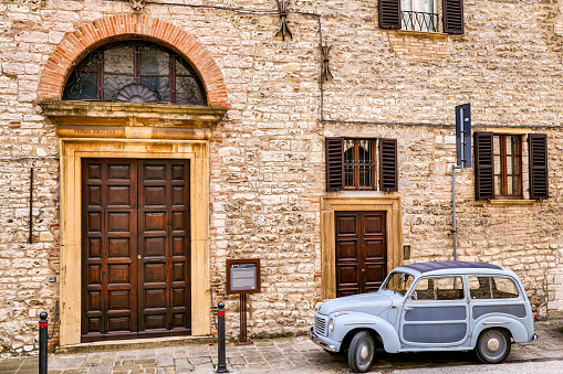 A vintage Fiat 500 C Giardiniera Belvedere (1953) parked in a suggestive stone alley on front the church of San Giovanni Decollato, built in 1508 by the Confraternity of Sant'Agostino, in the medieval town of Gubbio in Umbria, central Italy. An ancient city built on the communication routes between the Tyrrhenian coast and the Adriatic coast of Italy, an ally of Ancient Rome, Gubbio is famous for its artistic treasures and considered one of the most beautiful medieval villages in Italy. This Umbrian town is also known for the life of St. Francis of Assisi and the miracle of the encounter between him and a wolf, and for the spectacular Festa dei Ceri. The Umbria region, considered the green lung of Italy for its wooded mountains, is characterized by a perfect integration between nature and the presence of man, in a context of environmental sustainability and healthy life. In addition to its immense artistic and historical heritage, Umbria is famous for its food and wine production and for the quality of the olive oil produced in these lands. Image in high definition format.