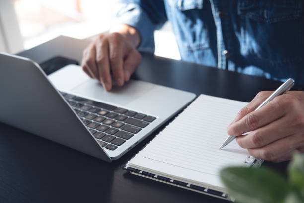 istock Close up of man working on laptop computer and hand with a pen writing on notebook on office table 1424224762