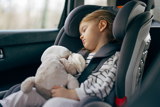Cute little girl in a child safety seat napping with her toy in the car. She is strapped in the seat and her head has fallen on the side.